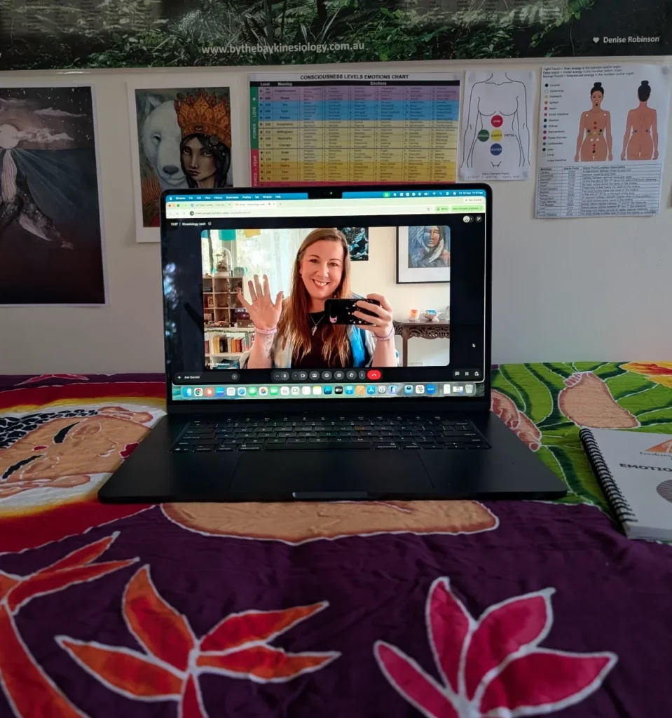 Kath smiling and waving on a laptop video call, with the laptop resting on a colourful treatment table beneath kinesiology charts.