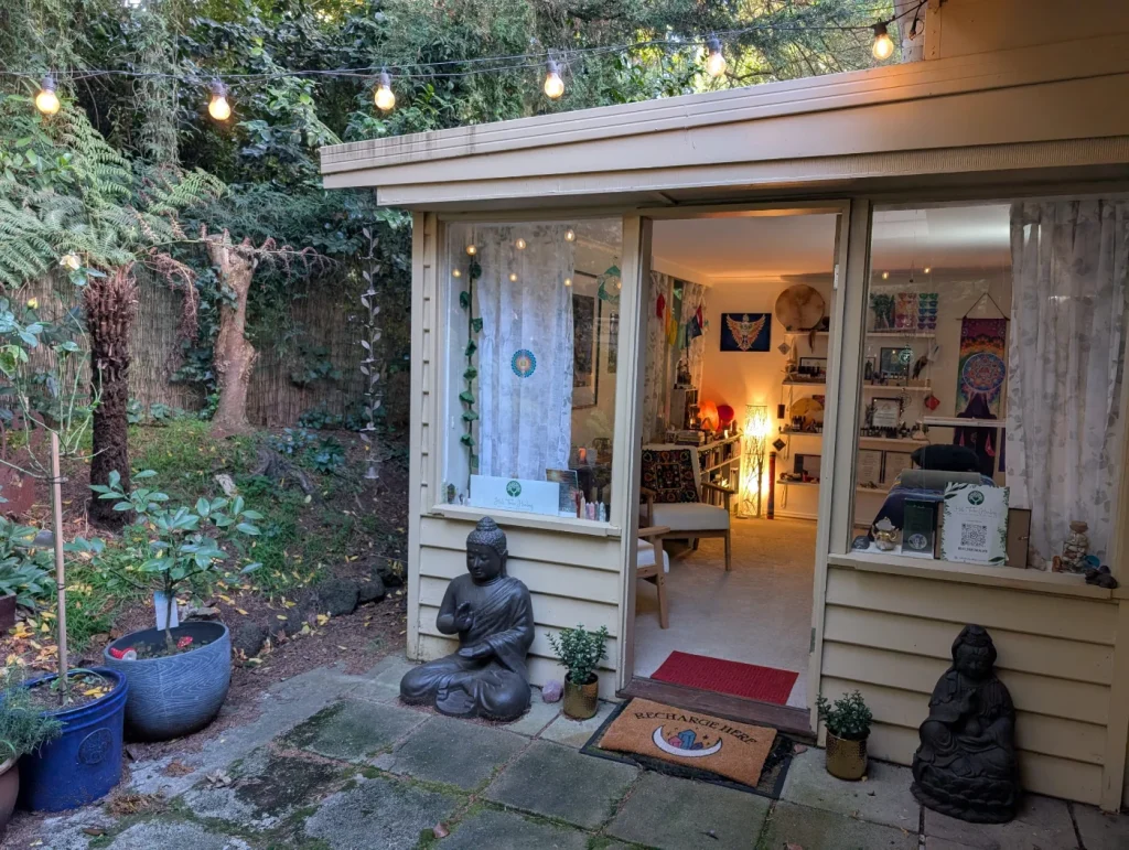 Open doorway to Hill Tribe Healing at The Patch, surrounded by garden greenery, string lights, potted plants and Buddha statues.
