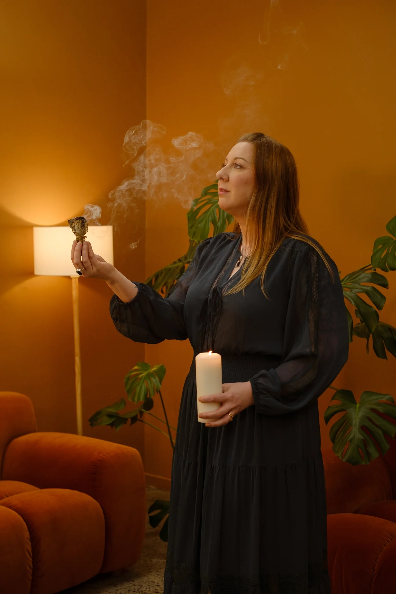 Kath holding a smoking smudge bundle and a lit pillar candle in a warmly lit room, with leafy plants.
